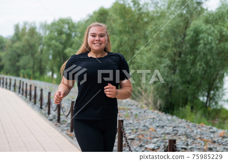 An obese young woman jogging outdoors. Fat beautiful smiling girl in a black tracksuit is engaged in fitness for weight loss on the waterfront. A woman runs on a summer day. An obese young woman jogging outdoors. Fat beautiful smiling girl in a black tracksuit is engaged in fitness for weight loss on the waterfront. A woman runs on a summer day. 75985229