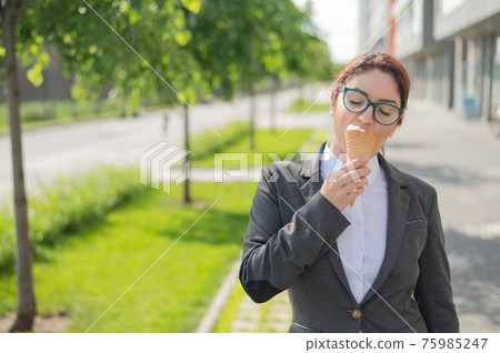 Business woman walks down the street and enjoys ice-cream. Happy girl in a suit eats an ice cream cone on a hot sunny summer day. Lunch break of an office employee. Business woman walks down the street and enjoys ice-cream. Happy girl in a suit eats an ice cream cone on a hot sunny summer day. Lunch break of an office employee. 75985247