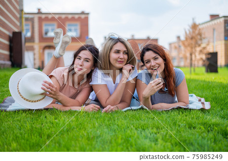 Three beautiful Caucasian young women lie on the lawn in the countryside. The blonde brunette and the redhead girls are resting in the park on the green grass Three beautiful Caucasian young women lie on the lawn in the countryside. The blonde brunette and the redhead girls are resting in the park on the green grass 75985249