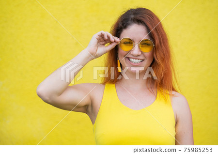 Portrait of a happy woman in sunglasses on a yellow background. Red-haired carefree girl in a dress and earrings tassels of the same color. Lady with an impeccable dazzling smile. Summer photo. Portrait of a happy woman in sunglasses on a yellow background. Red-haired carefree girl in a dress and earrings tassels of the same color. Lady with an impeccable dazzling smile. Summer photo. 75985253
