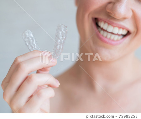 Close-up of orthodontic silicone transparent teeth aligner in female hands. A woman with a perfect charm smile holds a removable night retainer. Bracket for teeth whitening. Cropped photo. Close-up of orthodontic silicone transparent teeth aligner in female hands. A woman with a perfect charm smile holds a removable night retainer. Bracket for teeth whitening. Cropped photo. 75985255