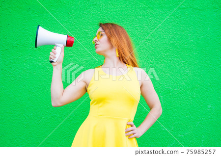Red-haired woman in sunglasses and a yellow dress holds a megaphone on a green background. Red-haired woman in sunglasses and a yellow dress holds a megaphone on a green background. 75985257