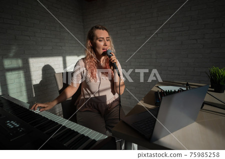 A female blogger sings and plays the live electronic piano. Portrait of a girl recording a song on a web camera and composing on a synthesizer. Online music lessons. Distance learning in quarantine. A female blogger sings and plays the live electronic piano. Portrait of a girl recording a song on a web camera and composing on a synthesizer. Online music lessons. Distance learning in quarantine. 75985258