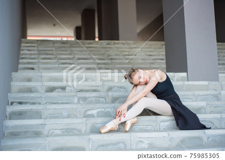 Ballerina in tutu posing sitting on the stairs A beautiful young woman in a black dress and pointe shoes is resting sitting in an elegant pose on the stairs from the outside 75985305