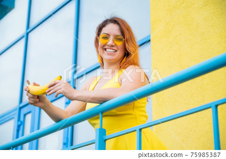 Red-haired Caucasian woman in sunglasses and yellow dress holding a banana outdoors. Red-haired Caucasian woman in sunglasses and yellow dress holding a banana outdoors. 75985387