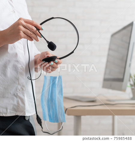 A faceless female employee holds a headset and a face mask. Woman with headphones and a medical mask in her hands 75985488