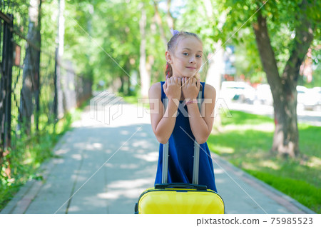 A contented little girl in a blue dress is standing alone in the street with a yellow suitcase and waiting for her parents. A child is going on a summer vacation trip. 75985523