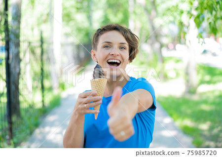 Charming Caucasian woman with a short haircut smiles and eats an ice cream cone and shows a thumb. Emotional girl with a beautiful smile enjoys gelato walking in the park. 75985720