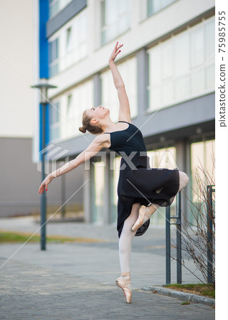Ballerina in a tutu posing against the backdrop of a residential building. Beautiful young woman in black dress and pointe shoes dancing ballet outside. Ballerina in a tutu posing against the backdrop of a residential building. Beautiful young woman in black dress and pointe shoes dancing ballet outside. 75985755