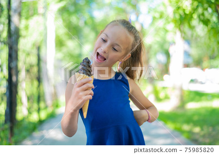 Happy little caucasian girl in blue dress eats chocolate ice cream cone outdoors. An emotional excited child enjoys a cooling gelato on a hot summer day. 75985820