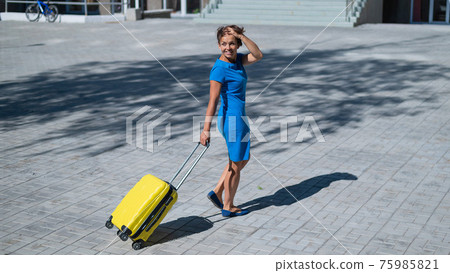 Happy woman walking down the street holding yellow suitcase with wheels 75985821