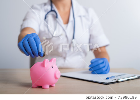 A faceless female doctor puts a coin in a piggy bank A faceless female doctor puts a coin in a piggy bank 75986189