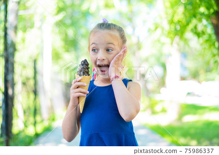 Happy little caucasian girl in blue dress eats chocolate ice cream cone outdoors. Emotional excited child enjoying a cooling gelato on a hot summer day in the park. 75986337