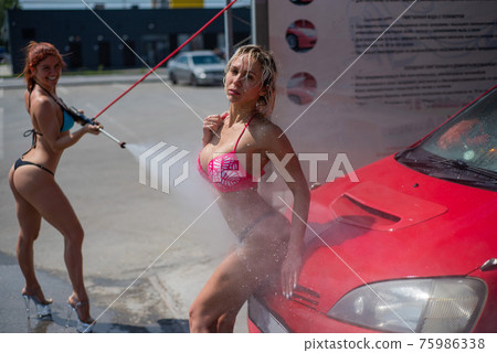 Luxury girls in seductive bikinis wash a car in a street car wash and water each other. Blonde and red-haired with beautiful figures wash a red car at a manual car wash. Self-care, dry cleaning salon. 75986338