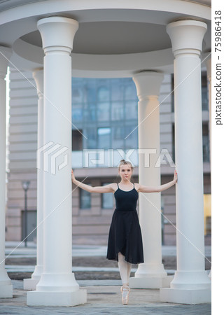 Ballerina in a black dress is a beautiful pose. A beautiful young woman in a black bodysuit is dancing in an elegant classical dance on a background of columns. 75986418