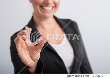 The woman is smiling and holding a transparent plastic aligner for bite correction 75987282