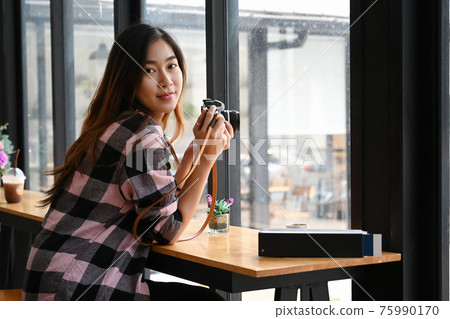 Young female photographer holding her camera and sitting near window in office. Young female photographer holding her camera and sitting near window in office. 75990170
