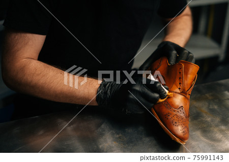Close-up hands of unrecognizable shoemaker in black gloves rubbing paint on light brown leather shoes with fingers. Close-up hands of unrecognizable shoemaker in black gloves rubbing paint on light brown leather shoes with fingers. 75991143