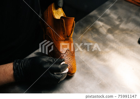 Close-up hands of shoemaker wearing black gloves rubbing paint on toe cup of light brown leather shoes with fingers. 75991147