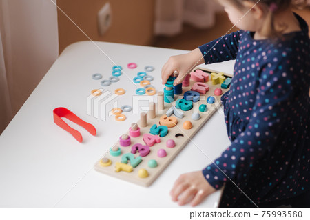 Closeup of kid's hand putting color rings on wooden column. Little girl play in educational game for children at the table. Wooden game with different colors and numbers. Intecactive game for clever Closeup of kid's hand putting color rings on wooden column. Little girl play in educational game for children at the table. Wooden game with different colors and numbers. Intecactive game for clever 75993580
