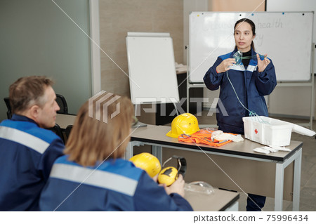 Young female worker demonstrating usage of respirator while standing by desk in front of colleagues 75996434