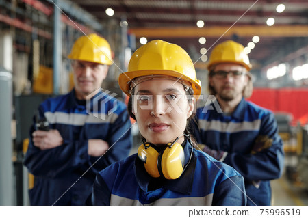 Young serious female worker of large industrial plant or factory in workwear and protective helmet Young serious female worker of large industrial plant or factory in workwear and protective helmet 75996519