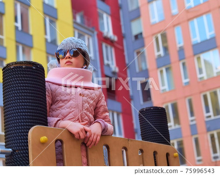 The girl 9 years old on the playground. The child is dressed in warm demi-season clothes a pink jacket and a grey hat. 75996543