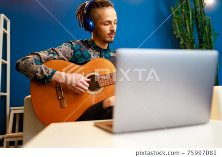 Young stylish caucasian man taking guitar class online at home using his laptop Young stylish caucasian man taking guitar class online at home using his laptop 75997081