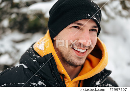 Young man in hoodie and warm jacket standing in a snowy park and smiling Young man in hoodie and warm jacket standing in a snowy park and smiling 75997507
