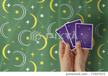 Hands of young woman with golden nail polish holding three tarot or oracle cards, on a green stars and crescent pattern table cloth. 75997635