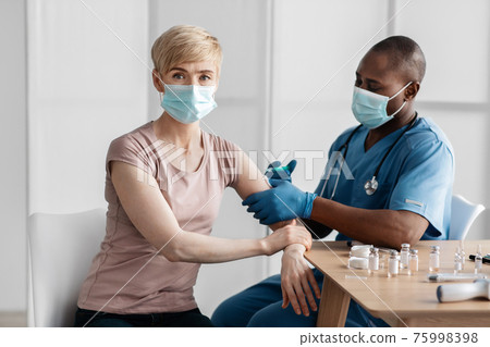 Adult african american lady sitting at doctor's office and getting modern Covid 19 shot injection in clinic Adult african american lady sitting at doctor's office and getting modern Covid 19 shot injection in clinic 75998398