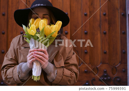 Beautiful girl in a raincoat riding a bicycle with with a vintage basket holds a bouquet of yellow Beautiful girl in a raincoat riding a bicycle with with a vintage basket holds a bouquet of yellow 75999391