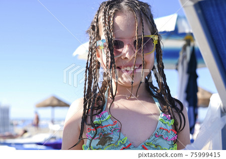 Little girl in a swimsuit on the beach plays, smiles. Child 7 years old on the equipped beach of Little girl in a swimsuit on the beach plays, smiles. Child 7 years old on the equipped beach of 75999415