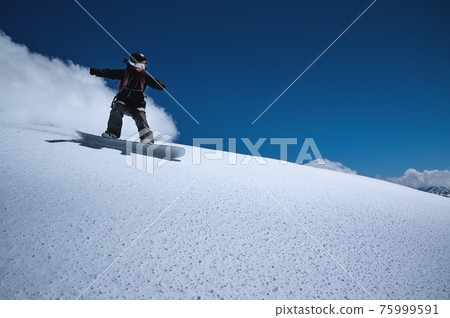 Young sportswoman woman on a snowboard rolls down a snowy slope against a blue sky on a sunny day Young sportswoman woman on a snowboard rolls down a snowy slope against a blue sky on a sunny day 75999591