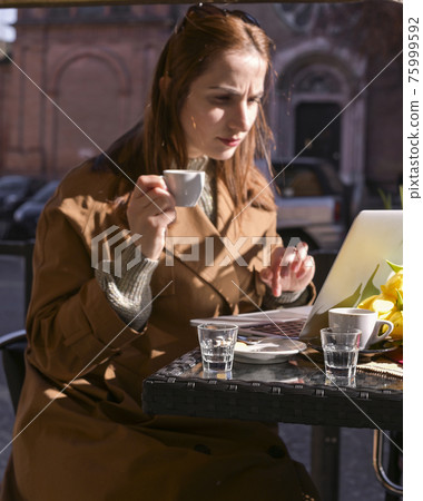 A young girl drinks coffee in a cafe on the street and works on the Internet on a laptop. Sunlight 75999592