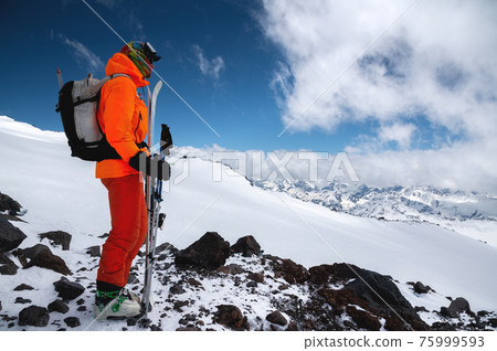 A young tall professional skier holds his skis while standing high in the mountains and looks at the snow-capped peaks. A young tall professional skier holds his skis while standing high in the mountains and looks at the snow-capped peaks. 75999593
