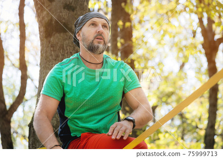 Portrait of an adult man with a beard in the autumn forest. Sportsman slackline next to a taut slackline Portrait of an adult man with a beard in the autumn forest. Sportsman slackline next to a taut slackline 75999713