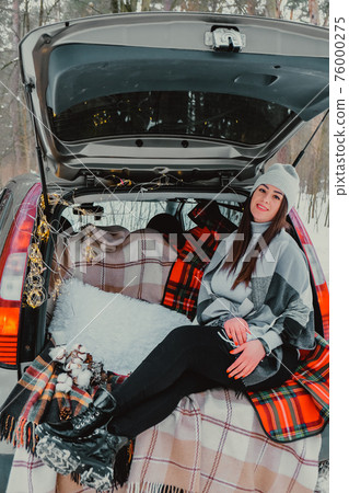 Brunette woman wrapped in blanket in trunk car. Travel in winter. Car decorated with festive Christmas lights. Outdoor picnic 76000275