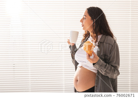Healthy beautiful pregnant woman drinking tea and eating croissant during lunch. Concept of high-calorie nutrition while waiting for birth of baby. Copy space. 76002671