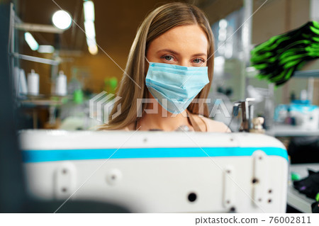 Young caucasian woman tailor working in sewing factory wearing protective medical mask Young caucasian woman tailor working in sewing factory wearing protective medical mask 76002811