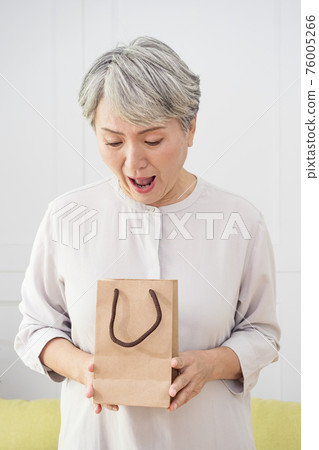 Portrait of an elderly Asian woman receives gift boxes, smiling and looking down while standing in a living room at home. 76005266