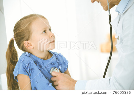 Doctor examining a child by stethoscope. Happy smiling girl patient dressed in blue dress is at usual medical inspection. Medicine concept 76005586