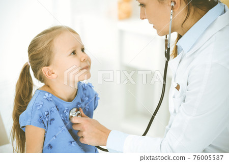 Doctor examining a child by stethoscope. Happy smiling girl patient dressed in blue dress is at usual medical inspection. Medicine concept 76005587