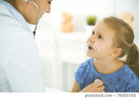 Doctor examining a child by stethoscope. Happy smiling girl patient dressed in blue dress is at usual medical inspection. Medicine concept 76005588