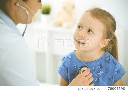 Doctor examining a child by stethoscope. Happy smiling girl patient dressed in blue dress is at usual medical inspection. Medicine concept 76005598