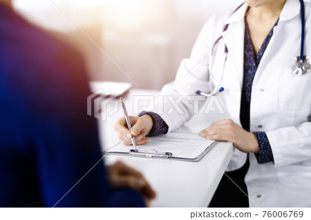 Unknown woman-doctor is writing some medical recommendations to her patient, while they are sitting together at the desk in the sunny cabinet in a clinic. Physician is using a clipboard, close-up Unknown woman-doctor is writing some medical recommendations to her patient, while they are sitting together at the desk in the sunny cabinet in a clinic. Physician is using a clipboard, close-up 76006769