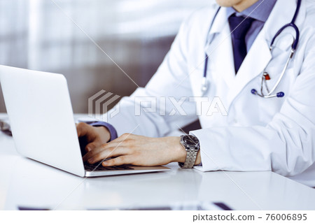 Unknown male doctor sitting and working with laptop in clinic at his working place, close-up. Young physician at work. Perfect medical service, medicine concept Unknown male doctor sitting and working with laptop in clinic at his working place, close-up. Young physician at work. Perfect medical service, medicine concept 76006895
