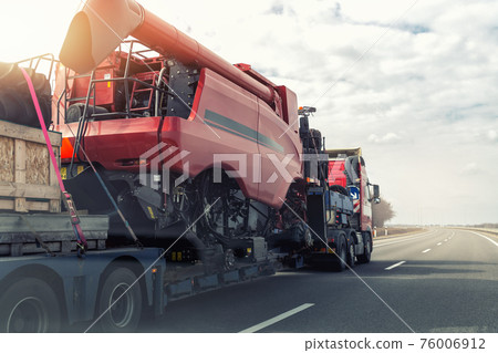 Heavy industrial truck with semi trailer platform transport disassembled combine harvester machine on common highway road on sunset or sunrise day. Agricultural equipment transportation service 76006912