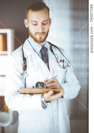 Friendly red-bearded doctor standing and writing with clipboard in sunny clinic. Medicine concept Friendly red-bearded doctor standing and writing with clipboard in sunny clinic. Medicine concept 76007599