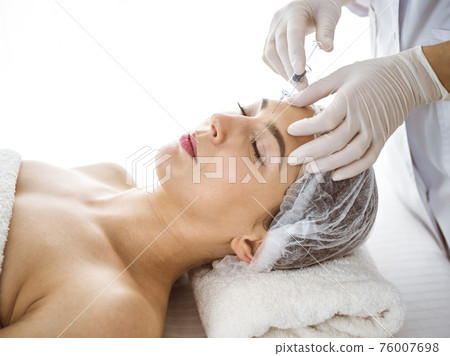 Beautician doing beauty procedure with syringe to face of young brunette woman in sunny clinic. Cosmetic medicine and surgery, beauty injections 76007698